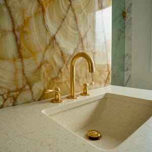 Könnte beinhalten: Eine goldfarbene Armatur und Armaturen sind auf einer hellen Arbeitsplatte mit einem eingebauten Waschbecken montiert. Der Hintergrund zeigt eine dekorative Wand mit einem marmorähnlichen Muster in Creme- und Goldtönen, was eine luxuriöse Badezimmerästhetik schafft.