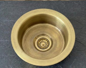 May include: A round, antique brass sink basin with a central drain. The basin has a smooth, brushed finish and a slightly flared rim. The drain features a decorative, star-shaped design. The sink is designed for kitchen or bathroom use.