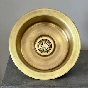 May include: A round, brass-colored kitchen sink with a central drain. The sink has a smooth, polished finish and a circular design. The drain is a darker brass color and has a decorative pattern. The sink is set against a neutral background.