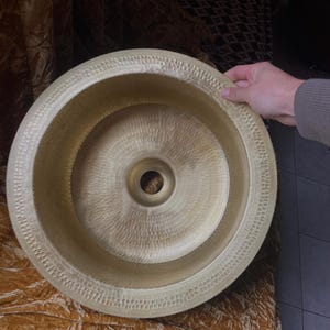 May include: A hammered brass sink with a circular design. The sink has a textured surface and a decorative rim. The center features a drain hole. The sink is a warm gold color and appears to be handmade.