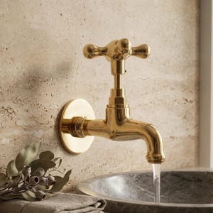 May include: A gold-colored wall-mounted faucet with a cross-shaped handle. The faucet is mounted on a circular base and has a curved spout. Water flows from the spout into a gray marble sink. The background is a textured beige wall.