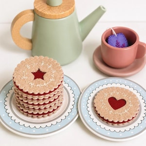 Puede incluir: Una tetera verde salvia con tapa de madera, una taza y plato rosa polvoriento con una bolsita de té de fieltro morada, y dos platos azul claro con lunares blancos. Un plato sostiene una pila de galletas de fieltro beige con estrellas, el otro un corazón.