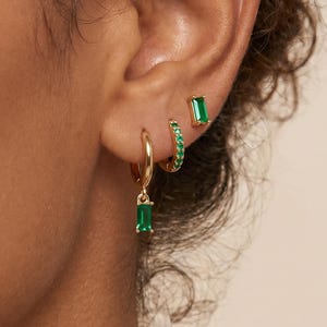 May include: Three gold earrings worn in one ear. The top earring is a small gold stud with a green baguette-cut stone. The middle earring is a small gold hoop with green stones. The bottom earring is a gold hoop with a green baguette-cut charm.
