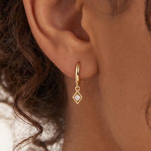 May include: A gold hoop earring featuring a small, diamond-shaped charm with a single clear stone at its center. The earring is worn in the earlobe, showcasing a polished metallic finish and a delicate, geometric design that hangs from the hoop.