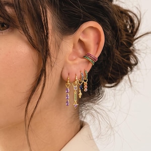Può includere: Un set di orecchini dorati e un ear cuff con pietre preziose colorate. L'ear cuff presenta pietre rosse e verdi, mentre gli orecchini pendenti includono varie forme, come gocce viola e piccoli accenti blu e verdi disposti in modo verticale.