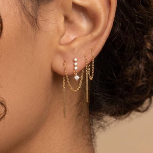 May include: Gold earrings with a dangling chain design. The earrings feature small, clear gemstones and a long, thin bar. The earrings are worn on the ear, with a focus on the jewelry.