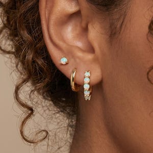 May include: A gold ear stack consisting of three pieces: a single opal stud earring in the upper lobe, a plain gold hoop in the middle, and a gold hoop earring featuring a row of five small, round, iridescent white opal gemstones.