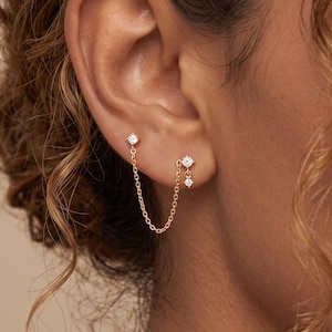 May include: A gold-toned double stud earring set connected by a delicate fine chain. Each stud features a small, clear, round-cut gemstone. One stud has a single gemstone, while the other features a gemstone with a small dangling stone below it.