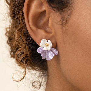 Könnte beinhalten: Ein Ohrstecker mit einem mehrschichtigen Blumendesign, bestehend aus einem kleinen weißen Blütenblatt oben und einem größeren, strukturierten, irisierenden hellvioletten Blütenblatt als Basis. Die Blume zeigt feine Adern und einen gelben Kern.