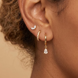 May include: A gold ear stud featuring two marquise-cut clear stones, a small gold hoop earring with clear stones, and a gold hoop earring with a dangling pear-shaped clear stone. These three gold earrings are worn in a single earlobe.