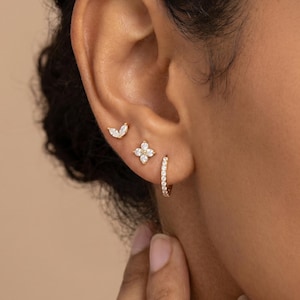 May include: A set of three gold earrings worn in a single ear. The top earring is a small marquise-cut stone stud, the middle is a four-petal flower stud with clear stones, and the bottom is a small gold hoop earring lined with tiny clear stones.
