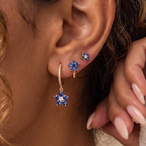 May include: A set of three gold-tone earrings featuring blue flower-shaped charms. One earring is a hoop with a dangling flower charm, while the other two are small studs shaped like flowers, all accented with small clear and blue stones.