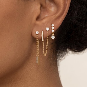 May include: A set of four gold-toned earrings worn in a single ear. The jewelry includes a stud with a dangling chain, a small hoop encrusted with clear stones, a stud connected to a chain, and a vertical drop earring featuring three round stones.