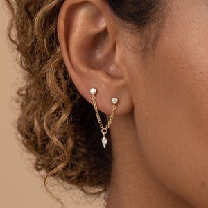 May include: A gold double-stud earring featuring two small round clear stones connected by a delicate gold chain that drapes downward, ending in a single pear-shaped clear stone drop. The earring is secured in two separate piercings on the earlobe.