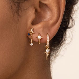 May include: Three gold-toned hoop earrings worn in a single ear. The first features a crescent moon with a blue stone and a dangling clear crystal. The second is a small hoop with a star-shaped crystal. The third is a hoop with a dangling star charm.