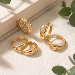 May include: Four gold hoop earrings featuring a polished finish and engraved sunburst motifs. Each sunburst design is centered with a small, clear, sparkling gemstone. The hoops are circular with a hinged closure mechanism, showcasing a consistent metallic yellow tone and intricate star-like detailing across the outer surface of each piece.
