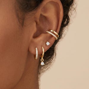 Könnte beinhalten: Eine Sammlung goldener Ohrringe und Ohrklemmen. Die Ohrringe umfassen kleine Creolen mit klaren Steinen, einen tropfenförmigen Ohrhänger und einen kleinen Ohrstecker. Die Ohrklemmen sind doppelt gebändert und mit klaren Steinen besetzt.