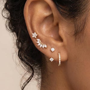 May include: A collection of gold earrings with clear gemstones. The earrings include a flower stud, a curved ear climber, a small square stud, a round stud, and a small hoop earring. The earrings are displayed on an ear.