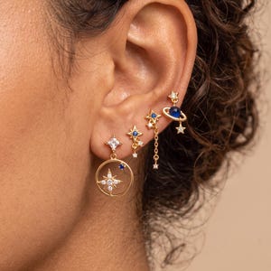 May include: A set of four gold-tone earrings featuring celestial motifs. The designs include a large circular charm with a central star, two small star-shaped studs with blue accents, and a planet-shaped charm with a blue sphere and dangling star.