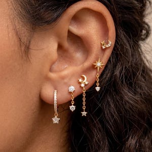 May include: A collection of gold-colored earrings, including a hoop with a star charm, a stud with a diamond, and dangling star and moon designs. The earrings are adorned with small, sparkling gems.