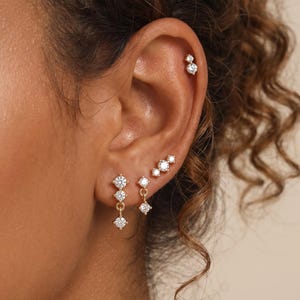 May include: A set of gold-toned earrings featuring clear, round-cut stones. The jewelry includes a vertical three-stone drop earring, a single-stone drop earring, a three-stone cluster stud, and a two-stone stud, all set in polished gold metal.