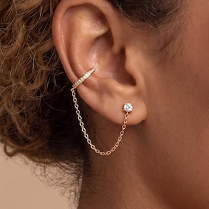 May include: A gold-toned ear cuff and stud earring set connected by a delicate gold chain. The ear cuff features a row of small, clear, sparkling stones, while the stud earring is a single, clear, round-cut stone set in a gold-toned metal base.