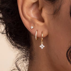 Könnte beinhalten: Ein Set aus goldenen Ohrringen, bestehend aus einem kleinen Stecker mit vierblättrigem Blumenmotiv, einem dünnen Goldring und einem Goldring mit einem hängenden vierblättrigen Blumenanhänger. Jedes Blumenelement ist mit kleinen klaren Steinen besetzt.