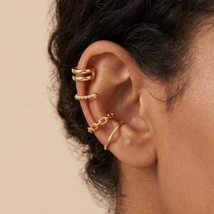 May include: A set of four gold-toned ear cuffs worn on the outer cartilage and conch of an ear. The collection includes a double-band cuff, a twisted rope-textured cuff, a small oval-link chain cuff, and a smooth, plain polished gold-toned band.