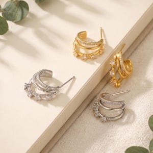 Könnte beinhalten: Vier mehrlagige Creolen mit dreireihigem Design. Zwei Ohrringe sind goldfarben und zwei sind silberfarben. Jeder Ohrring hat ein Band, das mit kleinen klaren Steinen besetzt ist, und ein weiteres Band mit größeren runden klaren Steinen.