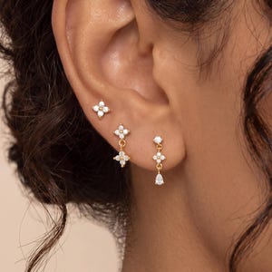 May include: Three gold-tone earrings worn in a single ear. The top earring is a four-petaled flower stud with clear stones. The middle earring is a flower stud with a dangling flower charm. The bottom earring is a stud with a dangling teardrop stone.