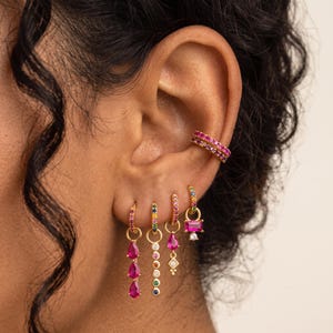 May include: A gold ear cuff with pink stones and four gold hoop earrings with dangling charms. The charms feature multi-colored stones, including pink pear-shaped drops, a vertical row of circular stones, a diamond-shaped accent, and a rectangular pink stone.