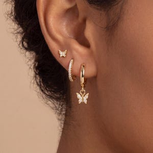 May include: A person's ear is shown with three gold earrings. There's a small butterfly stud, a small hoop with embedded crystals, and a larger hoop with a dangling butterfly charm, also with crystals. The earrings are gold in colour.