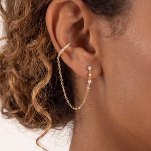 May include: A gold ear cuff and earring set connected by a delicate gold chain. The ear cuff features a row of small clear stones, while the earring consists of a single stud with a vertical drop of three small clear stones linked by the chain.