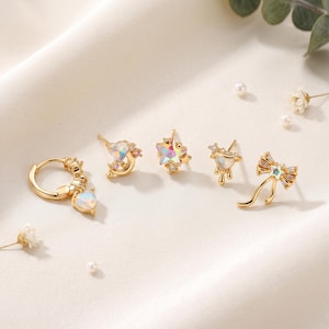 Può includere: Un set di cinque orecchini color oro con motivi iridescenti a forma di cuore, stella e fiocco. La collezione include un orecchino a cerchio con ciondolo a cuore, una mezzaluna con cuore, una stella, un cuore che cola e un fiocco, con cristalli.