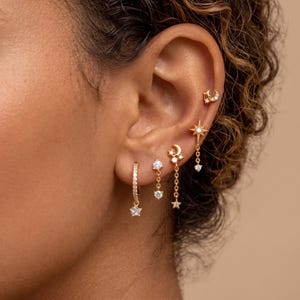 Peut inclure: Un ensemble de cinq boucles d'oreilles dorées aux motifs célestes. La collection comprend un petit clou étoile et croissant de lune, une pampille étoile, un croissant de lune avec pierres, une goutte de pierre et une créole avec breloque étoile.