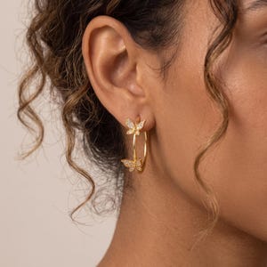 Peut inclure: Une boucle d'oreille créole en or ornée de deux petits papillons incrustés de pierres transparentes et scintillantes. Les papillons sont disposés le long du fin anneau circulaire en or, formant un design délicat et symétrique sur le lobe.