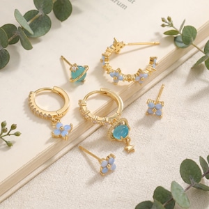 May include: A set of gold-tone earrings featuring celestial and floral motifs. The collection includes hoop earrings with small clear stones, studs shaped like light blue four-petaled flowers, and charms resembling a planet with a ring and a star.