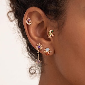 May include: A set of four gold-tone earrings featuring a crescent moon with a purple stone, a witch hat with a green stone, a spider web with a purple stone and a dangling spider, and a bat-winged figure with a white stone and orange base.