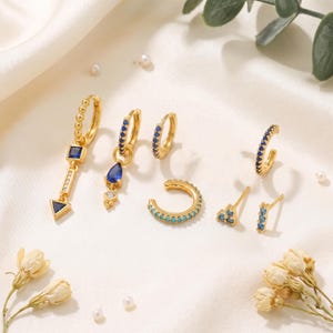 Puede incluir: Una colección de joyería en tono dorado que incluye pendientes de aro con detalles de piedras azules, un pendiente colgante en forma de flecha con una piedra cuadrada azul, un dije de piedra azul en forma de lágrima, un pendiente tipo ear cuff turquesa y pequeños pendientes de botón.