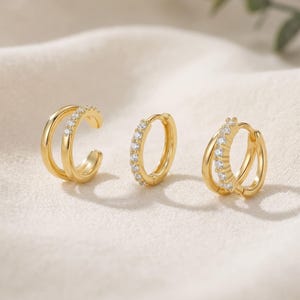 May include: Three gold-coloured earrings. One is a double-banded ear cuff with small, clear stones. The second is a small hoop earring with clear stones. The third is a double-banded hoop earring with clear stones. All earrings are on a cream-coloured surface.