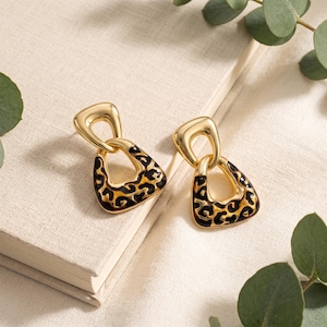 May include: A pair of gold-toned drop earrings featuring a polished metal top loop connected to a triangular base. The lower triangular section is filled with a black and brown leopard print pattern, creating a distinct contrast against the gold metal.