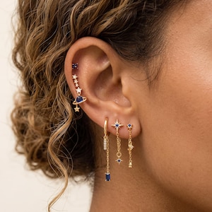 May include: A collection of gold-toned earrings featuring celestial motifs including stars and a planet with blue and clear stones. The set includes a multi-piercing climber earring, a hoop with a dangling baguette stone, and two chain-link drop earrings.