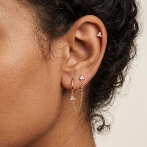 May include: A gold hoop earring with a delicate chain connecting to a small stud, featuring three tiny clear stones arranged in a cluster on both the hoop and the stud. An additional single stud with three clear stones is placed in the upper ear.