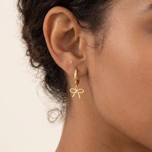 May include: A gold hoop earring featuring a small, dangling bow charm. The earring is constructed from a polished gold-tone metal, with a circular hoop that secures through the earlobe and a delicate bow pendant suspended from the bottom.