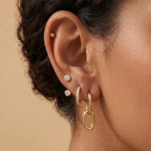 Könnte beinhalten: Ein goldener Ohrschmuck-Stapel bestehend aus einem kleinen Kugelstecker, zwei runden Ohrsteckern mit klaren Steinen, einem kleinen Creolen-Ohrring mit Pavé-Besatz und einem hängenden Ohrring mit ovalen Gliedern, angeordnet an Ohrläppchen und Knorpel.