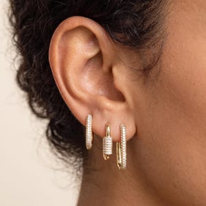 May include: Three gold-toned hoop earrings worn in a single earlobe. The first is a small pavé hoop, the second is a rectangular carabiner-style charm hoop with pavé stones, and the third is a longer, thin rectangular hoop with pavé detailing.