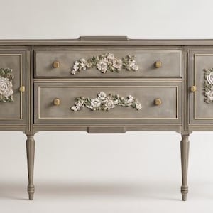 May include: A gray buffet with floral accents. The buffet has two drawers and two cabinet doors, each adorned with white and cream-colored floral designs. Gold-colored knobs and trim add detail to the piece. The buffet stands on four tapered legs.