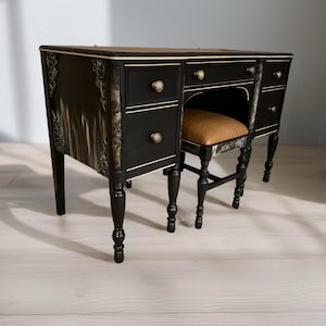 May include: A black vanity desk with a matching stool. The desk has three drawers on the left and two on the right, with gold-colored knobs. The stool has a brown cushion and a patterned seat back. The desk has gold trim.