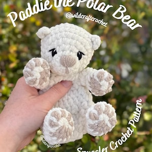 May include: A handmade, white crochet polar bear named "Paddie" with tan accents on the nose and paws. The bear is a "Snuggler Crochet Pattern" and is held in front of a blurred green background.