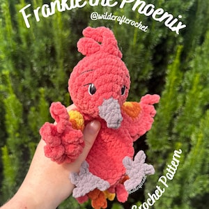 Peut inclure: Un jouet phénix crocheté à la main, rouge, avec un bec et des pieds gris, et des accents jaunes et orange. Le texte "Frankie the Phoenix" et "Snuggler Crochet Pattern" sont visibles. Le jouet est tenu dans une main sur un fond vert.